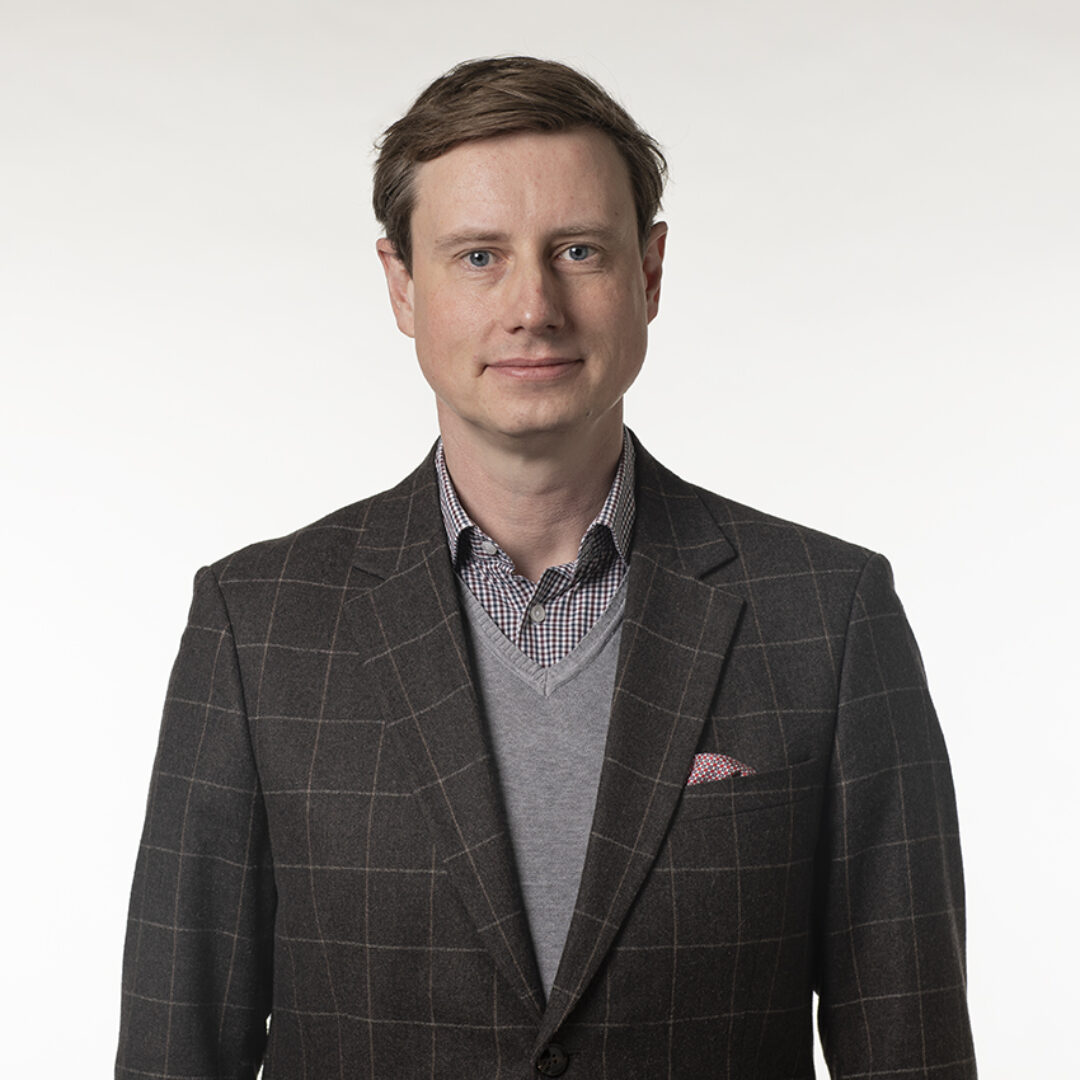 Prof. Dr. Sebastian Wolf trägt einen grauen Pullover und einen karierten Blazer. Er steht vor einem hellen Hintergrund und lächelt leicht.