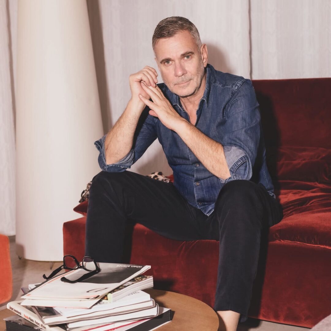 Wolfgang Benz sitzt auf einem roten Sofa, trägt ein blaues Hemd. Bücherstapel liegen auf einem Tisch.